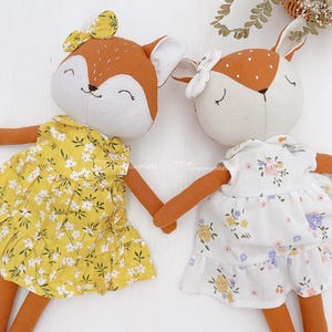 Pode incluir: Duas bonecas de pel&uacute;cia de raposa feitas &agrave; m&atilde;o vestindo vestidos florais. A raposa &agrave; esquerda est&aacute; vestindo um vestido amarelo com flores brancas e a raposa &agrave; direita est&aacute; vestindo um vestido branco com flores amarelas e roxas.