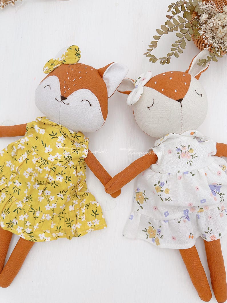 Pode incluir: Duas bonecas de pel&uacute;cia de raposa feitas &agrave; m&atilde;o vestindo vestidos florais. A raposa &agrave; esquerda est&aacute; vestindo um vestido amarelo com flores brancas e a raposa &agrave; direita est&aacute; vestindo um vestido branco com flores amarelas e roxas.