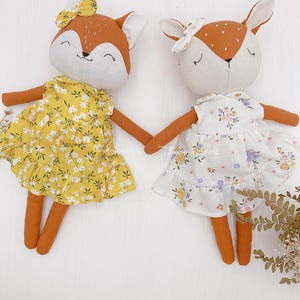 Pode incluir: Dois brinquedos de pel&uacute;cia feitos &agrave; m&atilde;o vestidos com vestidos de estampa floral. A raposa est&aacute; usando um vestido amarelo com flores brancas e a cor&ccedil;a est&aacute; usando um vestido branco com flores rosa e roxas.