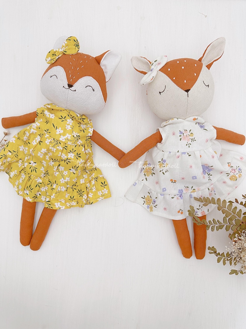 Pode incluir: Dois brinquedos de pel&uacute;cia feitos &agrave; m&atilde;o vestidos com vestidos de estampa floral. A raposa est&aacute; usando um vestido amarelo com flores brancas e a cor&ccedil;a est&aacute; usando um vestido branco com flores rosa e roxas.