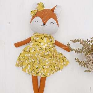 Pode incluir: Uma boneca de pel&uacute;cia de raposa feita &agrave; m&atilde;o vestindo um vestido amarelo floral com um la&ccedil;o branco na cabe&ccedil;a. A boneca tem um rosto sorridente com detalhes bordados.