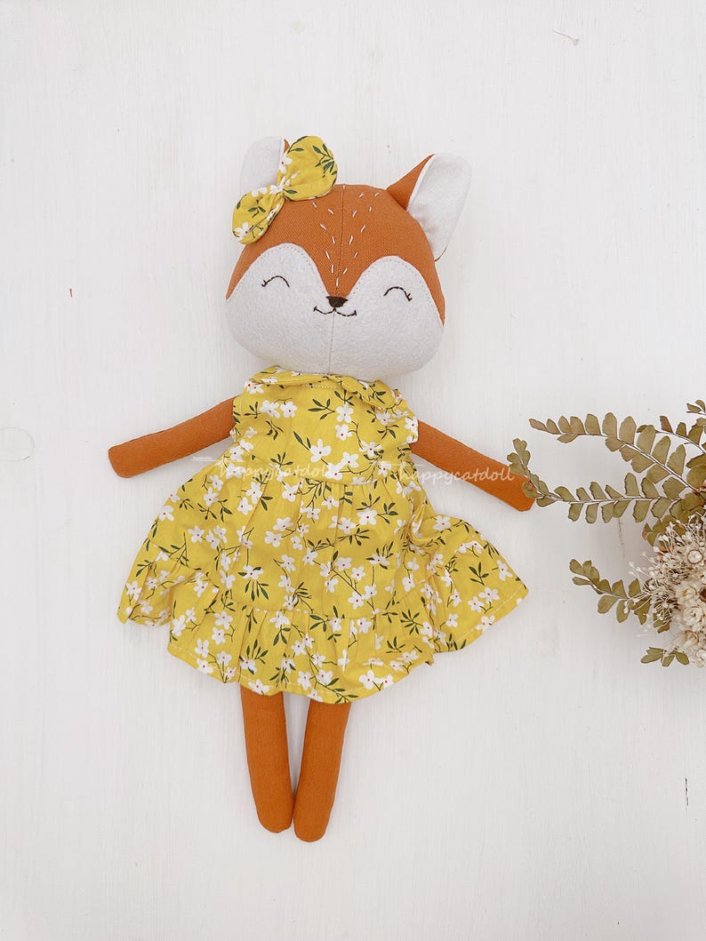 Pode incluir: Uma boneca de pel&uacute;cia de raposa feita &agrave; m&atilde;o vestindo um vestido amarelo floral com um la&ccedil;o branco na cabe&ccedil;a. A boneca tem um rosto sorridente com detalhes bordados.