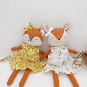 Pode incluir: Dois brinquedos de pel&uacute;cia feitos &agrave; m&atilde;o, uma raposa e um veado, ambos com vestidos florais. A raposa est&aacute; usando um vestido amarelo com flores brancas e o veado est&aacute; usando um vestido branco com flores rosa e roxas. Ambos os brinquedos t&ecirc;m pelo marrom e rostos bordados.