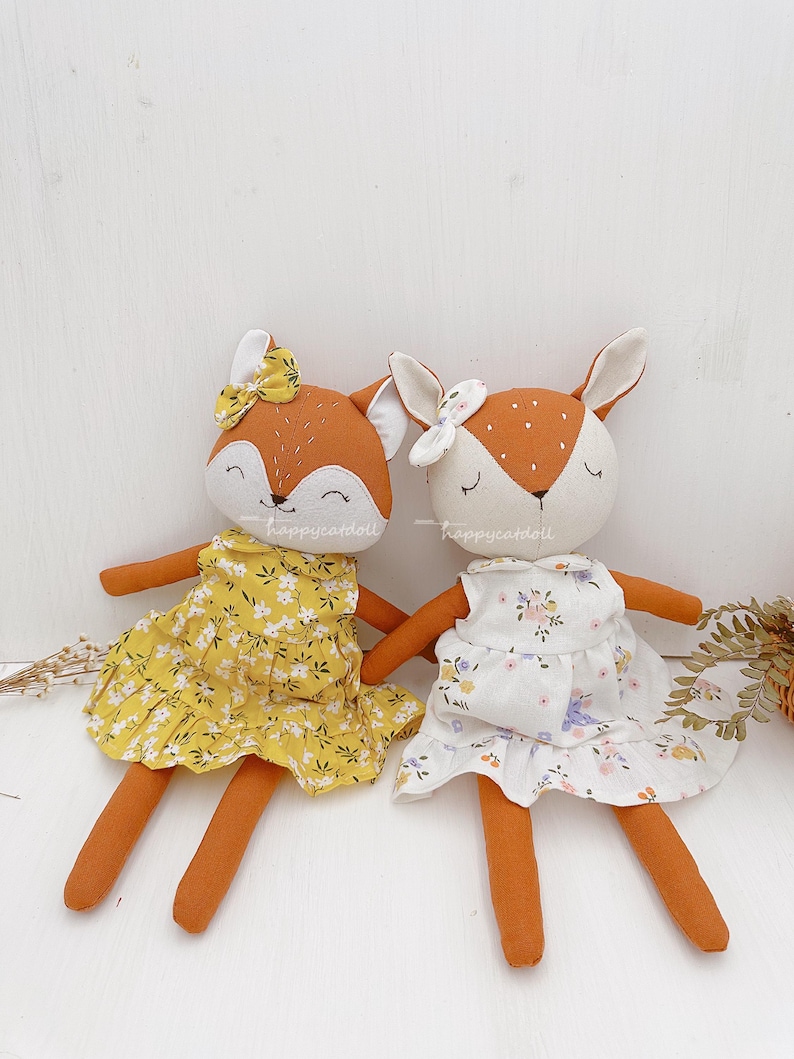 Pode incluir: Dois brinquedos de pel&uacute;cia feitos &agrave; m&atilde;o, uma raposa e um veado, ambos com vestidos florais. A raposa est&aacute; usando um vestido amarelo com flores brancas e o veado est&aacute; usando um vestido branco com flores rosa e roxas. Ambos os brinquedos t&ecirc;m pelo marrom e rostos bordados.