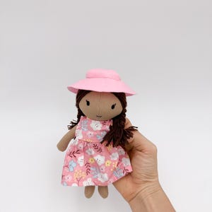Peut inclure: Poupée faite à la main avec des tresses brunes et un chapeau rose. La poupée porte une robe rose à motifs floraux. La poupée est en tissu et a un teint neutre.