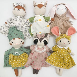 May include: A collection of six plush animal dolls featuring a fox, a deer, a rabbit, a cat, a raccoon, and another rabbit. Each doll is dressed in patterned fabric clothing, including floral dresses, aprons, and headbands in various colors.