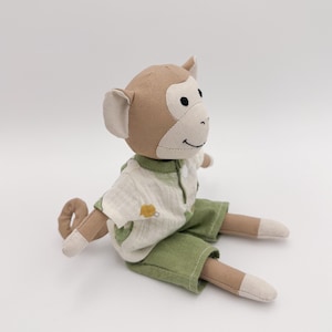 May include: A plush monkey doll with a tan head, ears, arms, and tail. The monkey wears a white and green shirt and green shorts. The doll is sitting on a white surface.