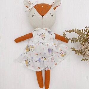 Pode incluir: Uma boneca de veado de pel&uacute;cia feita &agrave; m&atilde;o vestindo um vestido branco com padr&atilde;o floral. A boneca tem orelhas marrons, um nariz marrom e pernas marrons. A boneca est&aacute; usando uma faixa de cabe&ccedil;a rosa com um la&ccedil;o.