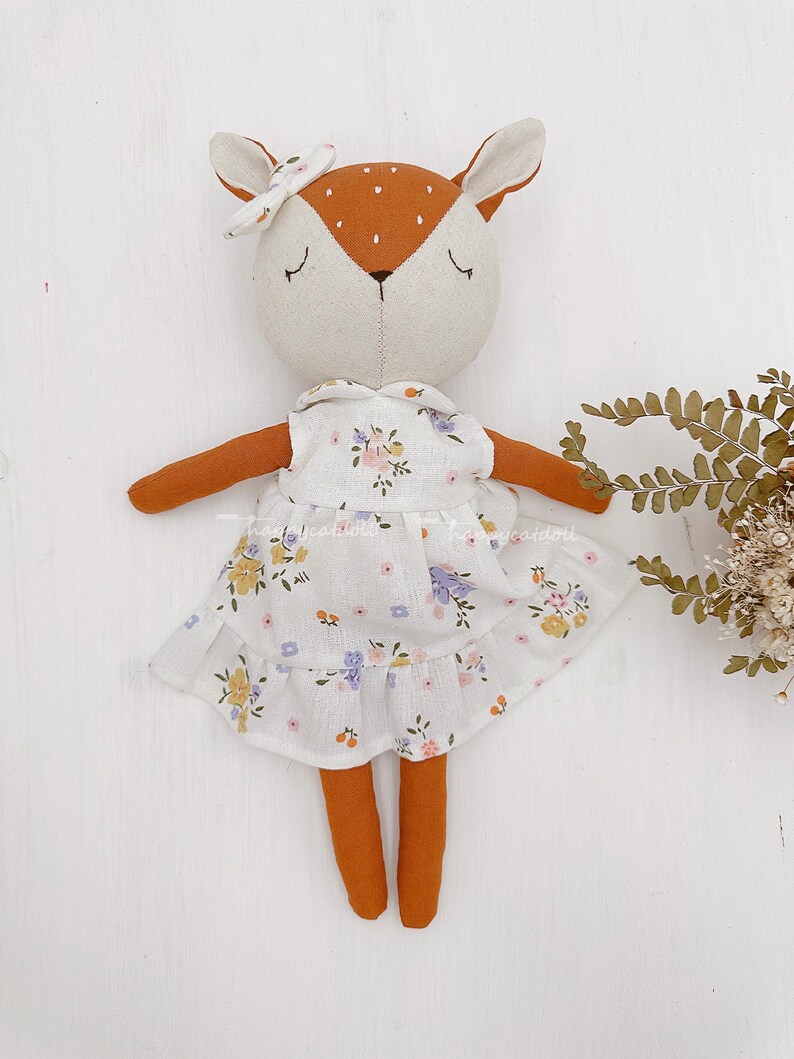 Pode incluir: Uma boneca de veado de pel&uacute;cia feita &agrave; m&atilde;o vestindo um vestido branco com padr&atilde;o floral. A boneca tem orelhas marrons, um nariz marrom e pernas marrons. A boneca est&aacute; usando uma faixa de cabe&ccedil;a rosa com um la&ccedil;o.