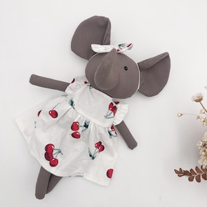 May include: A gray elephant plush toy wearing a white dress with a red cherry print. The elephant has large gray ears, a trunk, and a small bow on its head. The dress has a ruffled hem and short sleeves. The toy is on a white background.