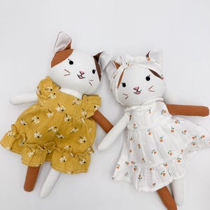 Pode incluir: Duas bonecas de gato de pelúcia com rostos bordados e orelhas castanhas. Uma boneca usa um vestido amarelo mostarda com um padrão floral branco e mangas com folhos. A outra usa um vestido branco com uma pequena estampa floral laranja e uma fita para o cabelo a combinar.