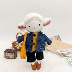 Può includere: Una bambola di agnello di peluche con giacca di jeans, camicia gialla, pantaloncini neri e scarpe marroni. L'agnello ha una testa bianca e soffice e orecchie foderate di rosa, con una borsa a tracolla gialla. La bambola è su una superficie bianca.