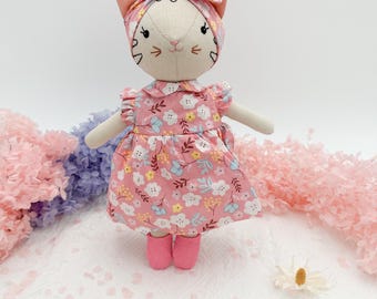 Könnte beinhalten: Handgefertigte Stoffpuppe in Form einer Katze, die ein rosa geblümtes Kleid, ein passendes Stirnband und rosa Stiefel trägt. Die Puppe hat gestickte Gesichtszüge und ausgestreckte Arme. Der Hintergrund umfasst rosa und violette Dekorationselemente.