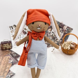 May include: A handmade stuffed bunny doll wearing an orange cap, bow tie, and a blue and white striped overall. The bunny has long brown ears with white lining and a matching orange shoulder bag. A charming, whimsical toy.