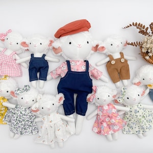 Peut inclure: Plusieurs poupées d'agneau faites à la main, chacune avec une tenue unique. Les poupées portent des robes, des salopettes et un béret de différentes couleurs et motifs. Un petit panier avec des fleurs séchées est également présent.
