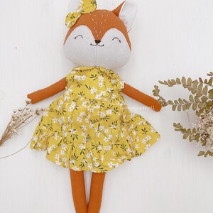 Pode incluir: Uma boneca de pel&uacute;cia de raposa vestindo um vestido amarelo com estampa floral branca. A boneca tem um corpo marrom, um rosto branco e um la&ccedil;o amarelo na cabe&ccedil;a.