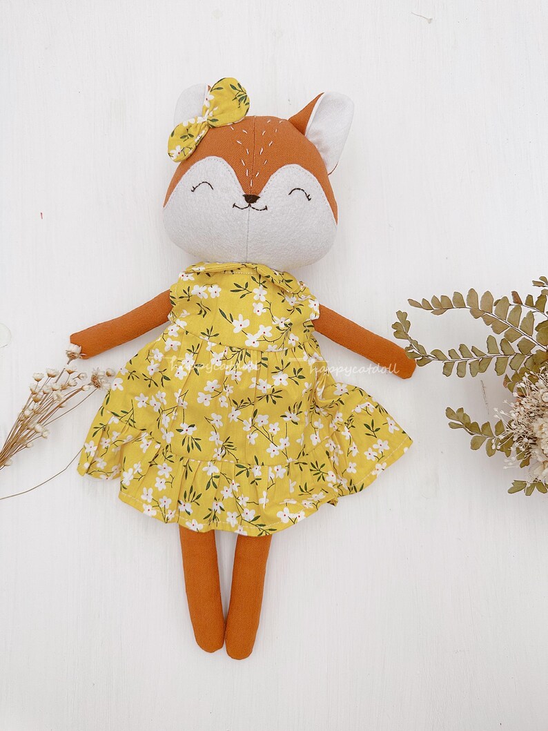 Pode incluir: Uma boneca de pel&uacute;cia de raposa vestindo um vestido amarelo com estampa floral branca. A boneca tem um corpo marrom, um rosto branco e um la&ccedil;o amarelo na cabe&ccedil;a.
