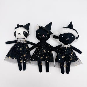 May include: Three handmade plush dolls: a panda, a cat, and a sheep, each wearing a black dress with gold star accents and a matching witch hat. The panda has a black bow, the cat has white ears, and the sheep has a white fluffy collar.