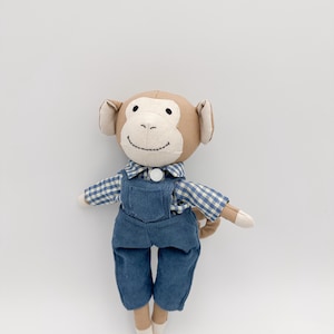 Puede incluir: Un muñeco de mono de peluche con cabeza, orejas y brazos color canela, y una cara sonriente. Viste una camisa a cuadros azul y blanco y un peto de pana azul. El muñeco tiene una pequeña cola y pies blancos.