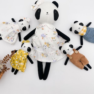 May include: A family of five panda plush toys. The mother panda is wearing a white dress with floral print and a pink bow. The other pandas are wearing different outfits, including a yellow dress, a blue and yellow outfit, and a brown overall.