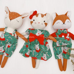 Op de afbeelding: Drie pluche speelgoedjes gekleed in kerstthema outfits. Een bruin hert met een overall, een witte kat met een rode jurk en een vos met een rood shirt en overall. Alle drie de speelgoedjes hebben een groene en rode rendier- en snoepstokprint.