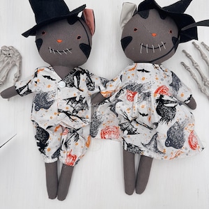 May include: Two handmade gray cat dolls dressed for Halloween. One wears a black top hat and pajamas, the other a witch's hat and dress. Both have stitched smiles and wear outfits with a Halloween print featuring bats and pumpkins.
