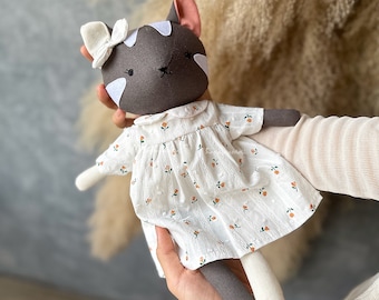 Op de afbeelding: Een grijze en witte kattenpop die een witte jurk met bloemenprint draagt. De pop heeft een witte strik op haar hoofd.
