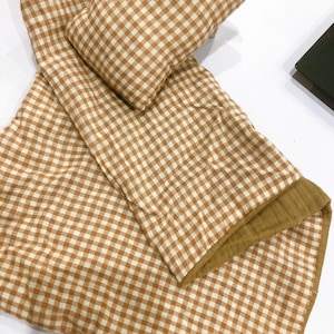 K&ouml;nnte beinhalten: Eine braune und wei&szlig;e Gingham-Babydecke mit einem braunen und wei&szlig;en Gingham-Kissen. Die Decke hat auf einer Seite eine weiche, flauschige Textur.