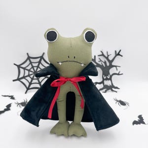 May include: A handmade Halloween-themed plush frog dressed as a vampire. The frog is olive green with large, round eyes and a black cape tied with a red bow. Decorative bats, spiders, and a spiderweb are in the background.