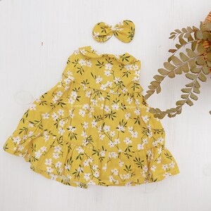Pode incluir: Um vestido amarelo com estampa floral branca e uma faixa de cabe&ccedil;a com la&ccedil;o amarelo combinando. O vestido tem uma bainha com babados e uma gola Peter Pan.