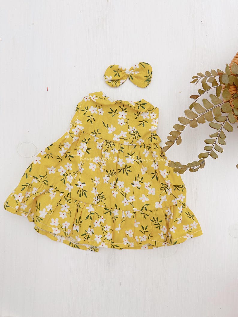Pode incluir: Um vestido amarelo com estampa floral branca e uma faixa de cabe&ccedil;a com la&ccedil;o amarelo combinando. O vestido tem uma bainha com babados e uma gola Peter Pan.