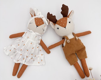 May include: Two handmade deer dolls. One doll wears a white dress with orange floral accents and a matching headband. The other doll wears brown corduroy overalls. Both dolls have brown antlers, arms, and legs, against a white background.