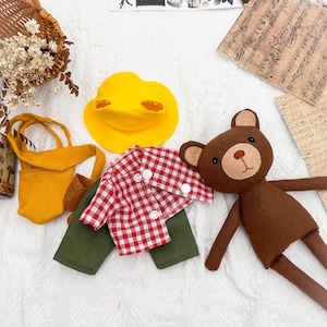 May include: A selection of handmade doll clothing and accessories. The image shows a red and white checked shirt, green trousers, a yellow duck hat, and a brown teddy bear doll. A yellow tote bag and a wicker basket are also visible.