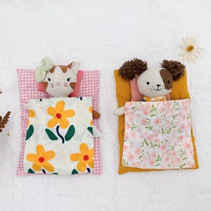 May include: Two plush toy animals, a cat and a dog, are nestled in miniature beds. The cat rests on a pink gingham pillow with a floral blanket. The dog lies on a yellow pillow with a floral blanket. Daisies are scattered around.