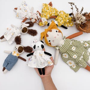 Peut inclure: Une collection de jouets en peluche faits à la main, comprenant un panda, un chat et deux chiens. Les jouets sont habillés de robes à fleurs et de salopettes. Les jouets sont disposés sur un fond blanc.