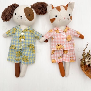 Puede incluir: Dos juguetes de peluche, un perro y un gato, que llevan pijamas a juego con un estampado de caritas sonrientes amarillas. El perro lleva un pijama azul y blanco, y el gato lleva un pijama rosa y blanco.