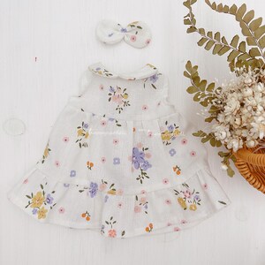 Pode incluir: Um vestido branco sem mangas com estampa floral em tons de rosa, amarelo e roxo. O vestido tem uma bainha com babados e uma gola Peter Pan. Uma faixa de cabe&ccedil;a branca com la&ccedil;o est&aacute; inclu&iacute;da.