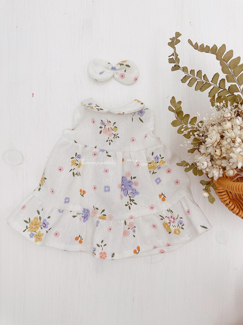 Pode incluir: Um vestido branco sem mangas com estampa floral em tons de rosa, amarelo e roxo. O vestido tem uma bainha com babados e uma gola Peter Pan. Uma faixa de cabe&ccedil;a branca com la&ccedil;o est&aacute; inclu&iacute;da.