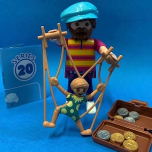 Puede incluir: Un juego de figuras Playmobil Serie 20 con un titiritero con un títere, una maleta con monedas y una caja con el número 20. El titiritero lleva un gorro azul y una camisa a rayas. El títere es verde con lunares amarillos.