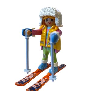 May include: A Playmobil skier figure, wearing a yellow vest, pink and blue striped shirt, green trousers, and a white fur hat. The figure is holding blue ski poles and standing on orange skis with blue and white designs.