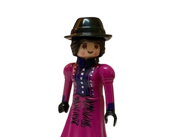 May include: A Playmobil figure of a woman in a long, magenta dress with black accents and a black hat. The figure has a painted face and black shoes. The dress has a decorative design on the front.