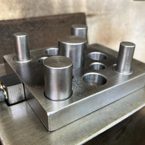 May include: A square metal plate featuring a grid of circular holes and several upright cylindrical metal posts. A small black rectangular block with two silver-colored screws is attached to the side of the plate, which rests on a larger metal base.