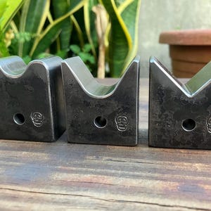 May include: Three dark metal V-shaped objects with a skull emblem. Each object has a hole and a curved top. The objects are arranged on a wooden surface, with green foliage in the background.