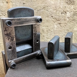 Può includere: Un utensile in metallo con un telaio rettangolare e un componente scorrevole, insieme a due pezzi di metallo abbinati. L'utensile ha un aspetto industriale usurato, con bulloni visibili e una finitura scura e invecchiata. Gli oggetti sono su una superficie di legno.