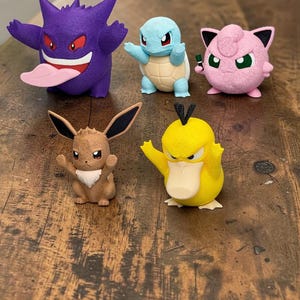 May include: A collection of five colorful Pokémon figurines. The figures include Gengar, Squirtle, Jigglypuff, Eevee, and Psyduck. The figurines are displayed on a wooden surface.
