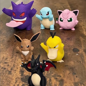 May include: A collection of six small 3D printed figures featuring a purple Gengar with a pink tongue, a light blue Squirtle, a pink Jigglypuff, a brown Eevee, a yellow Psyduck, and a black Charizard with red wings and a cream-colored belly.
