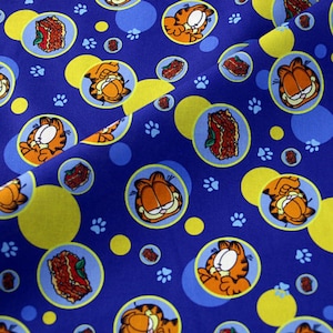 Puede incluir: Tela azul con un patrón repetido del gato Garfield, lasaña y huellas de patas. El diseño incluye círculos amarillos y azules. El gato de dibujos animados está en varias poses.