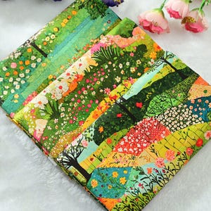 May include: A stack of colourful fabric squares with a vibrant floral and landscape design. The fabric features a patchwork of green, orange, and blue sections with trees, flowers, and foliage. Pink flowers are visible in the background.