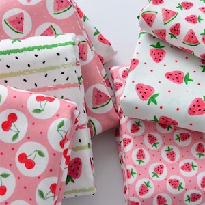 Puede incluir: Primer plano de varias piezas de tela dobladas con estampados de frutas. Las telas presentan dise&ntilde;os de cerezas, fresas y rodajas de sand&iacute;a en rosa, blanco y verde. Las telas est&aacute;n dispuestas en una pila, mostrando la variedad de patrones.