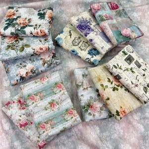 May include: A collection of folded fabric squares featuring various floral patterns, including pink and peach roses, blue accents, and vintage-style script text. The textiles display diverse botanical prints, muted color palettes, and intricate illustrations on soft, patterned surfaces.